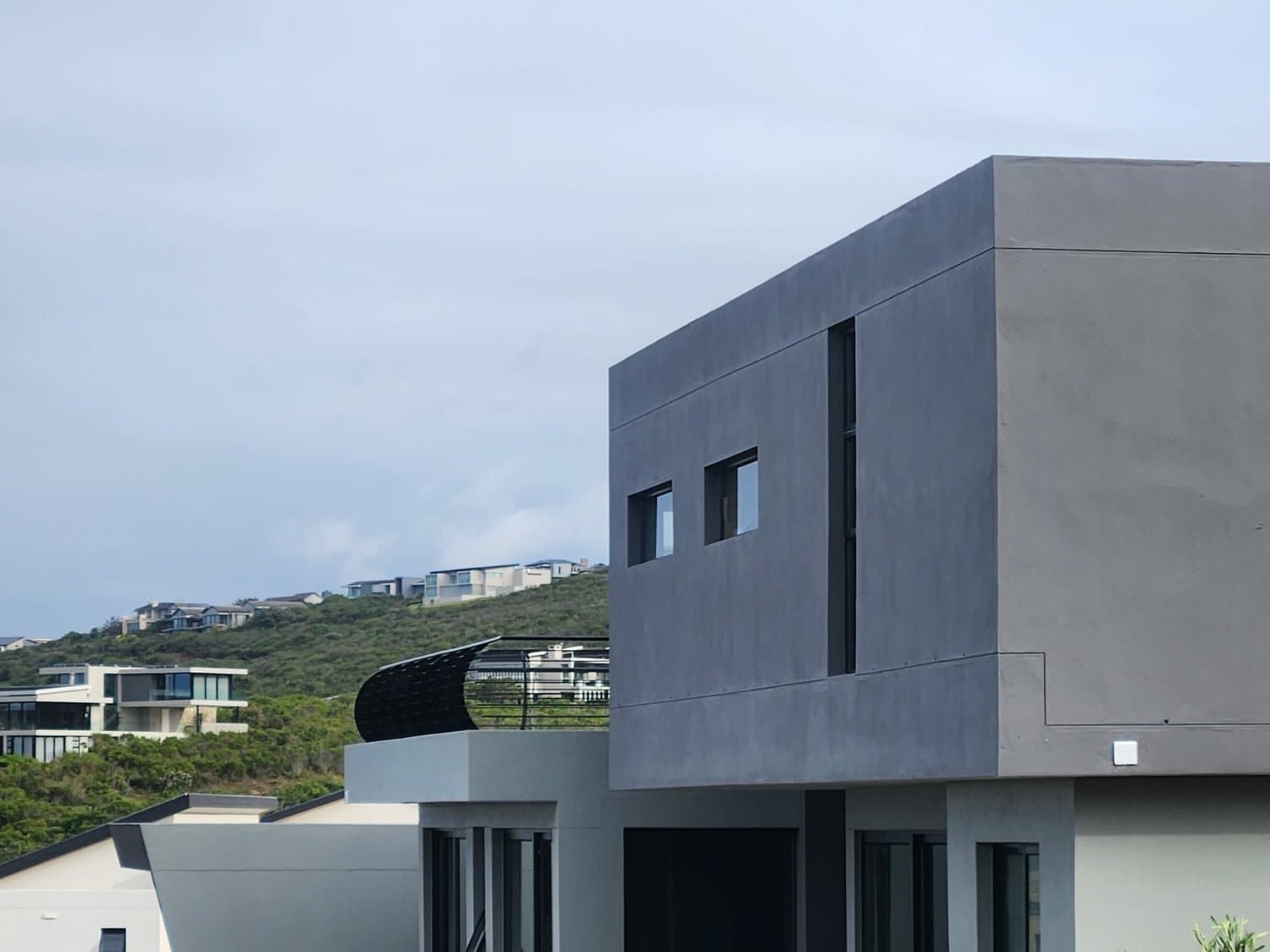 A completed home in charcoal render with a flat roof, above a wooded slope