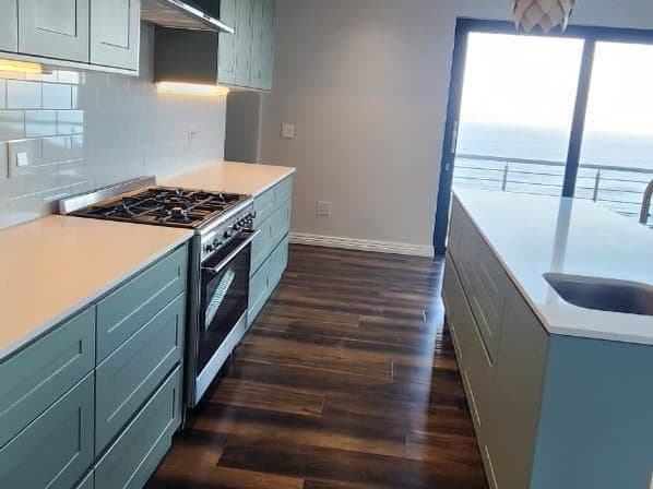 A fitted kitchen in deep teal with a gas hob, timber floors and a sea view