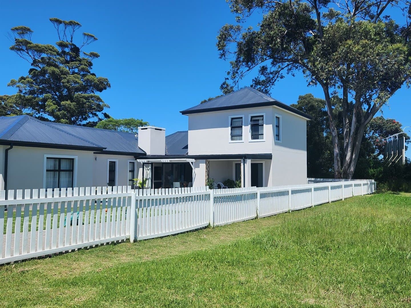 A finished two storey home behind a white picket fence, under tall gum trees